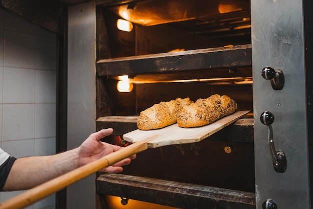 Анонимный мужчина достает хлеб с семенами лопатой из духовки в пекарне