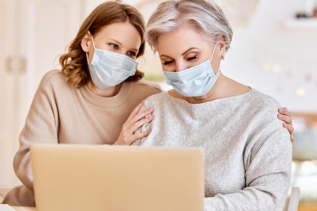 Anonymous serious young woman in casual clothes and face mask\
pointing at screen of laptop while helping elderly mother working\
remotely at home during coronavirus pandemic