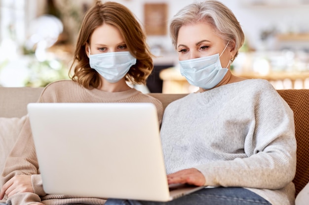 Anonymous serious young woman in casual clothes and face mask\
pointing at screen of laptop while helping elderly mother working\
remotely at home during coronavirus pandemic