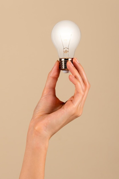 anonymous young slim female with slender fingers holding aloft light bulb with filament in display in hand against brown background