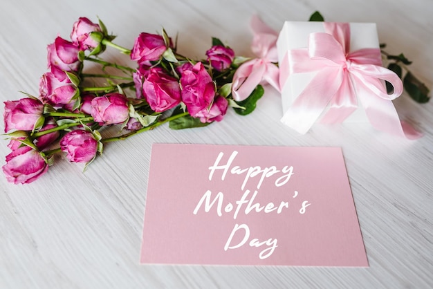 Ansichtkaart met boeket van kleine roze rozen geschenkdoos op witte rustieke houten achtergrond happy mother's day tekst teken wenskaart met bloemen concept vakantie wenskaart