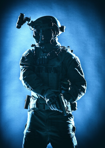 Anti terrorist squad fighter, army elite forces soldier in mask, with night vision device and tactical radio headset on helmet, armed service pistol standing ready for action, low key studio shoot