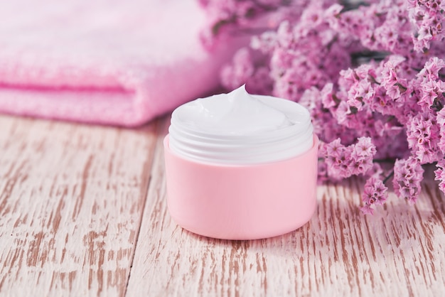 Crema antirughe antietà per la cura del corpo o per la cura del viso igiene idratante lozione con fiori rosa in barattolo di plastica con asciugamano sul tavolo di legno. vasetto di plastica rosa per crema per pelli sensibili.