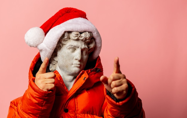 Antique statue dressed in down jacket and Santa Claus hat on pink wall
