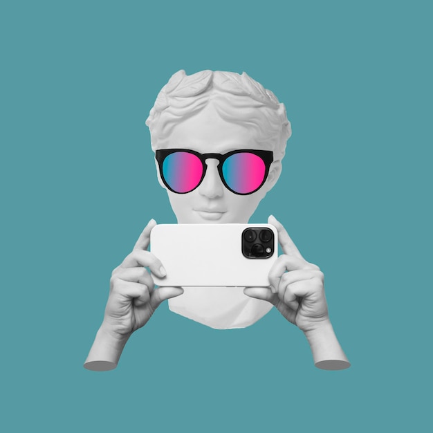 Photo antique statue's head in sunglasses holding mobile phone with photo camera in hands taking picture