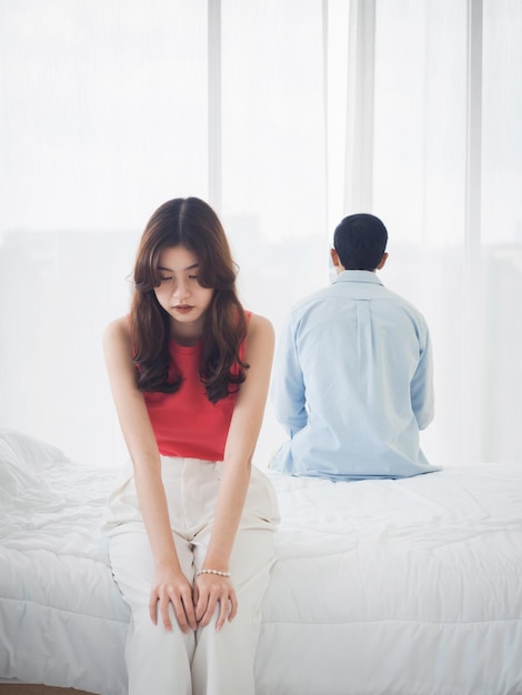 The anxiety of asian couple lovers on the bed sad young woman\
and man sitting on the bed with relationship difficulties feeling\
sad and thinking in the bedroom at home near the glass window