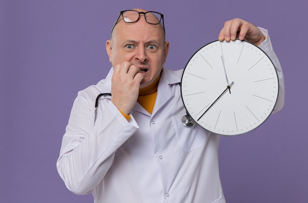 Anxious adult man with glasses in doctor uniform with stethoscope holding clock and biting his nails