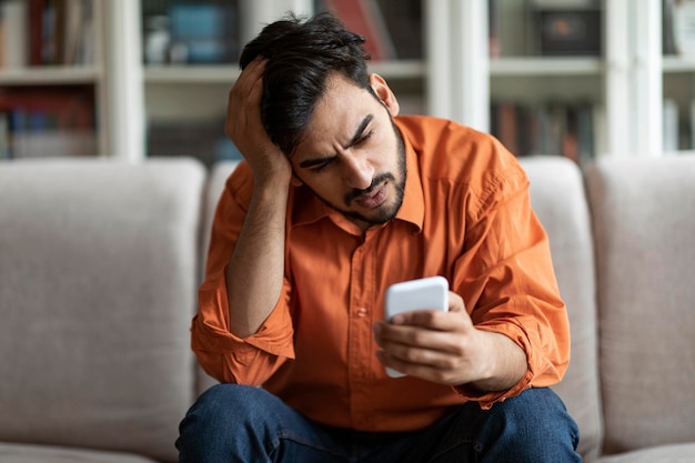 Anxious arab guy sitting on couch at home holding smartphone