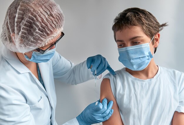 注射器が怖い、顔を作る不安な子供。医者、医者、看護師、白いガウンとフェイスマスクの医療従事者が10代の少年にワクチンを接種します。新しい通常のコンセプト、両方の人がフェイスマスクを着用