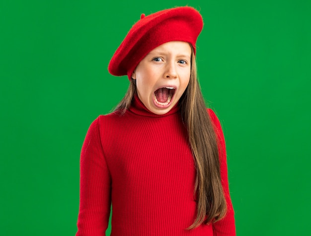 Ansiosa bambina bionda che indossa un berretto rosso che guarda la telecamera e urla isolata sulla parete verde con spazio per le copie