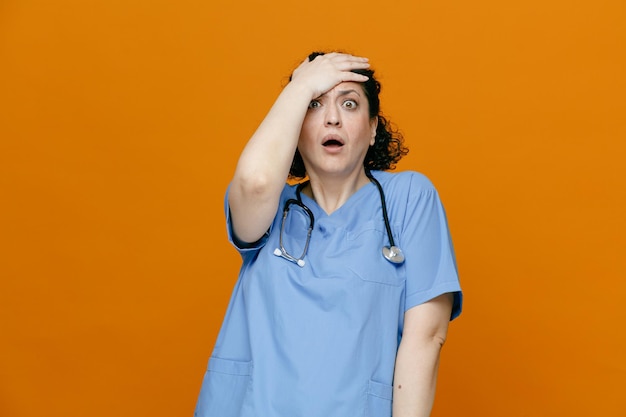 Anxious middleaged female doctor wearing uniform and stethoscope around her neck keeping hand on forehead looking at camera isolated on orange background