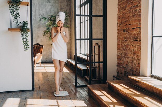 Anxious slim caucasian woman wrapped in towel taking step onto scale to measure her weight