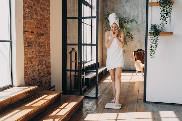 Anxious slim caucasian woman wrapped in towel taking step onto scale to measure her weight