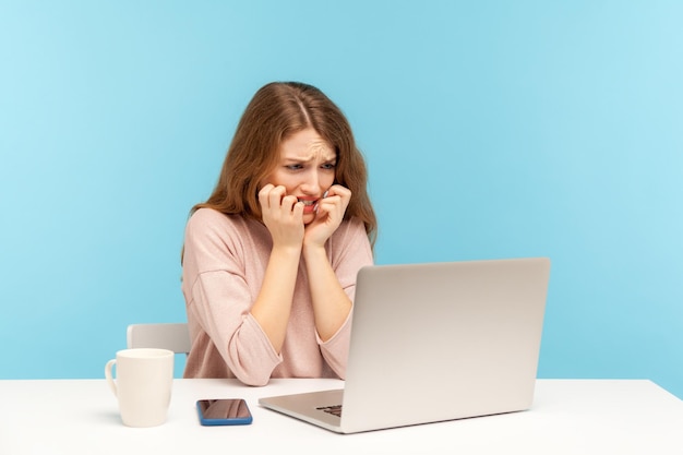 Anxious upset young woman employee sitting at workplace looking at laptop screen with nervous worry expression and biting nails scared of software error job deadline indoor studio shot isolated