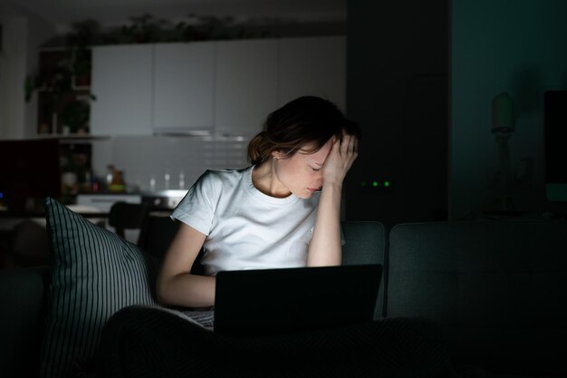 Anxious woman sitting on couch using laptop at night read sudden bad news and cover face with palm