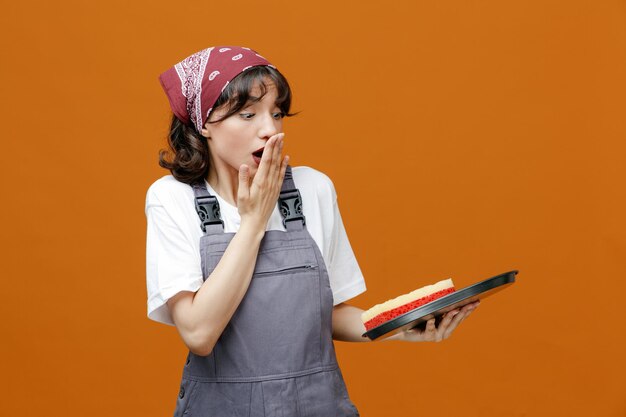 Photo anxious young female cleaner wearing uniform and bandana holding sponge in tray keeping hand on mouth looking at sponge isolated on orange background