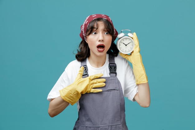 Anxious young female cleaner wearing uniform bandana and rubber gloves holding alarm clock near head looking at side while keeping hand on chest isolated on blue background