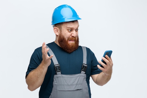 불안한 젊은 남성 건설 노동자는 안전 헬멧과 유니폼을 입고 흰색 배경에 고립 된 공기에 손을 유지하고 휴대 전화를보고 찾고