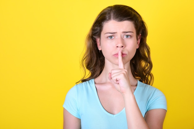 Anxious young woman presses a finger to her lips on a yellow wall