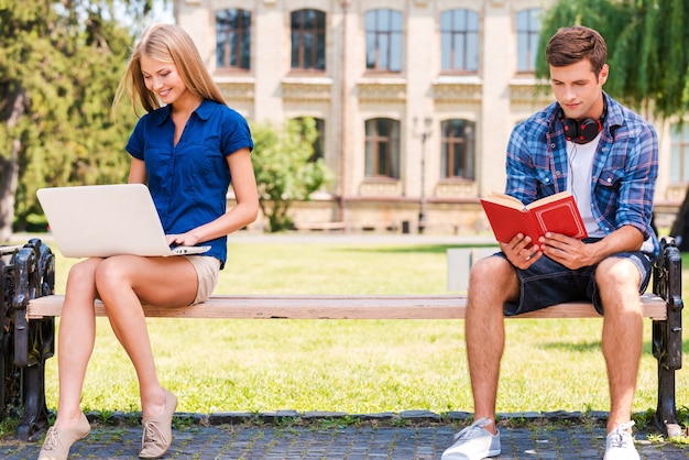 Apart ontspannen. knappe jonge man die op de bank zit en een boek leest terwijl een mooie vrouw in de buurt van hem zit en de computer gebruikt