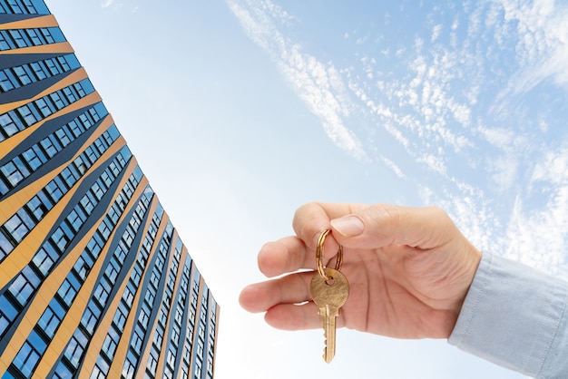 Apartment key in a man's hand. Brass house door lock key. Modern building against the blue sky. View from below.