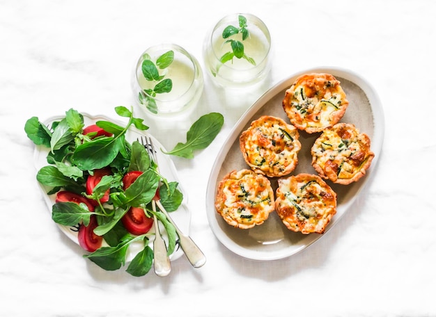 Aperitif table mini quiche with salmon and zucchini fresh tomato arugula salad and mint cocktail on a light background top view