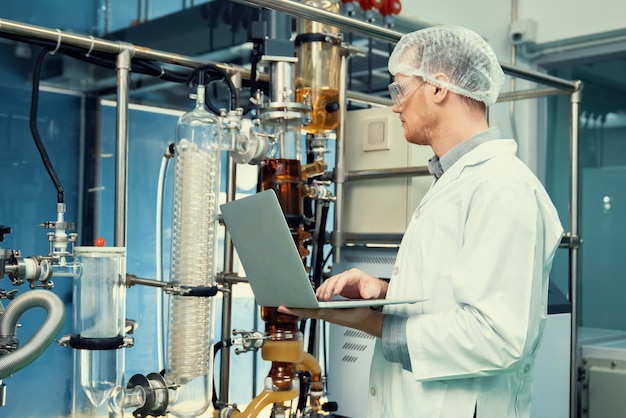 Apothecary scientist working with laptop near CBD oil extractor in laboratory