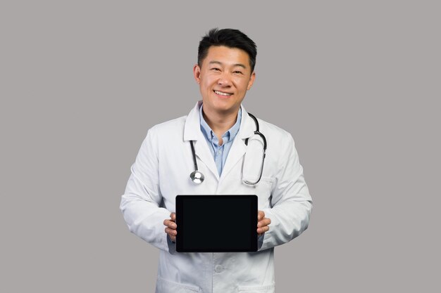 App for health care remote Smiling korean chinese male therapist in white coat with stethoscope shows tablet