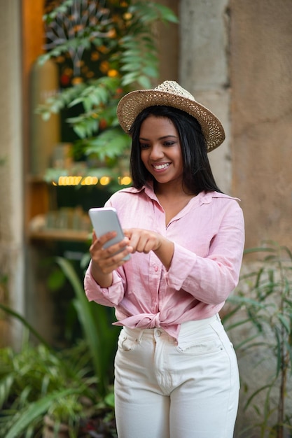 週末のオンラインアプリのアプリは、帽子をかぶったスタイリッシュな若いミレニアル世代の黒人女性観光客が電話を使用することを嬉しく思います