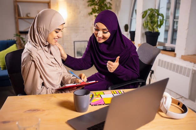 Apparaten, techniek. Gelukkige en jonge twee moslimvrouwen thuis tijdens de les, studeren in de buurt van computer, online onderwijs. Cultuur, tradities, moderne mensen. Kijken op pc-monitor, winkelen of praten.