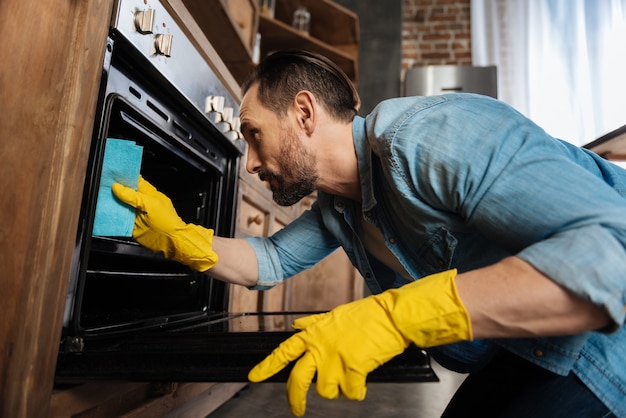 Appealing bearded male cleaner leaning over oven while cleaning it and wearing gloves