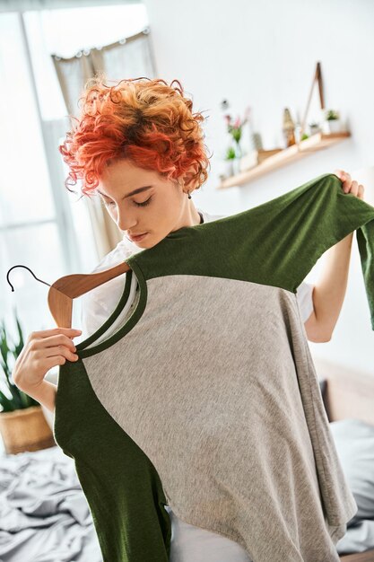 appealing red haired extravagant person picking up stylish clothes near rack at home leisure time