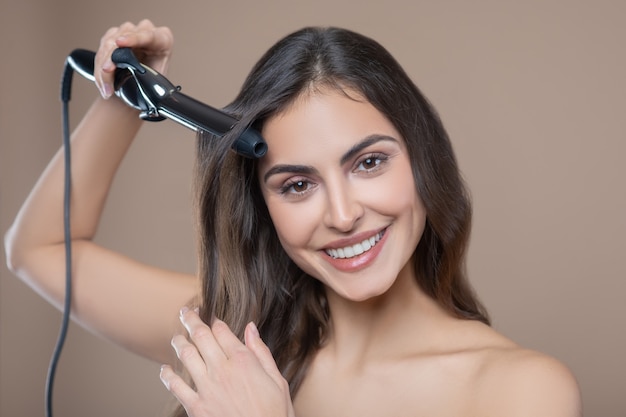 Appearance, hairstyle. Happy woman with long dark hair doing her hair with curling iron on beige background