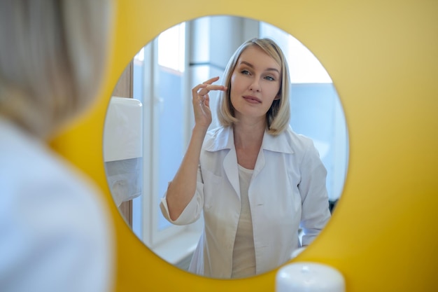 Appearance. Young pretty blonde dosctor in a lab coat standing at the mirror