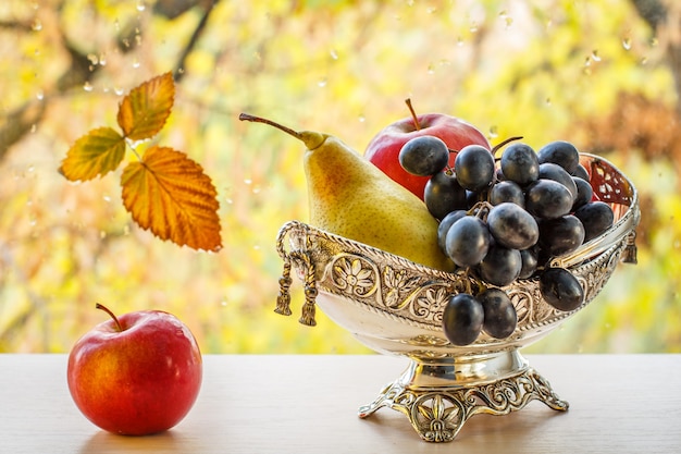 Appel en metalen vaas met gele peer en tros druiven. Droog blad op vensterglas met waterdruppels en herfstbomen op de achtergrond.