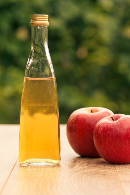 Appelazijn in glazen fles met metalen dop en verse rode appels op houten planken met groene natuurlijke achtergrond. Biologische voeding voor de gezondheid