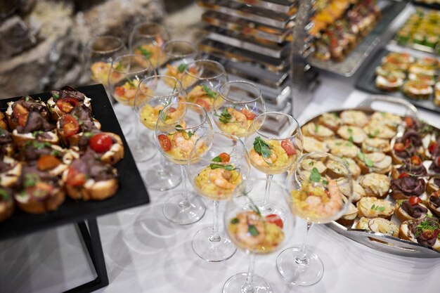 Foto appetiserende snacks van zeevruchten, vlees, groenten en fruit op borden en onderborden op een wit tafelkleedje versierd met een boeket bloemen tegen een stenen muurachtergrond tijdens een banket