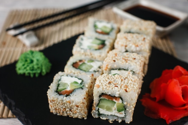 Appetising fresh california sushi roll set with salmon in sesame served on black slate with ginger and wasabi, close up. Japanese healthy seafood.