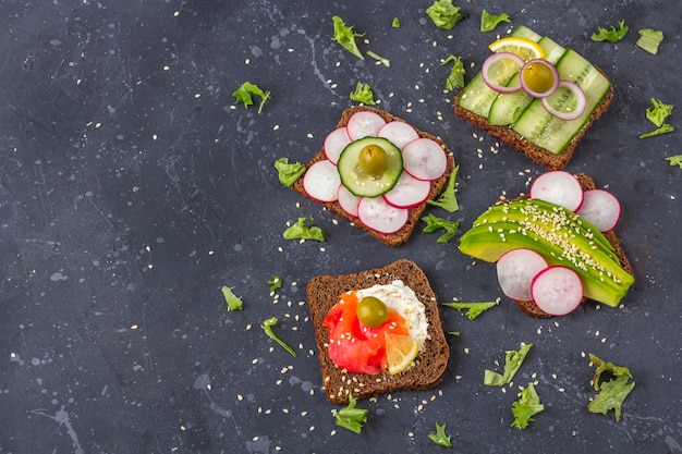 Appetizer, open sandwich with different toppings: salmon and vegetables (avocado, cucumber, radish) on dark background. Healthy eating