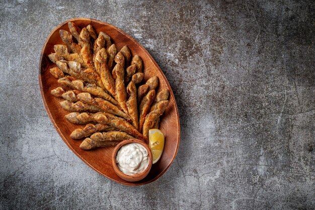 An appetizer in a restaurant, fried sprat on a wooden plate with lemon and cream sauce against a gray stone table