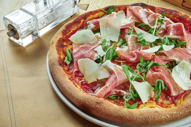 Appetizing baked pizza with prosciutto di parma, parmesan and arugula with crispy crust on a wooden table. Restaurant table setting. Close up, selective focus