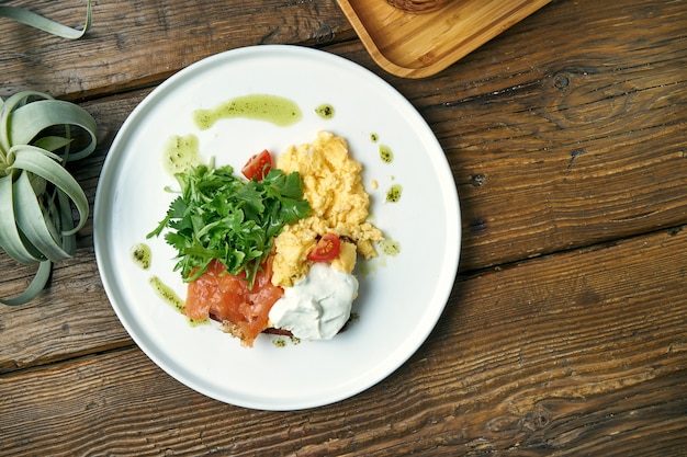 Colazione appetitosa - uova strapazzate, con rucola, salmone e pane tostato con yogurt greco in un piatto su un legno