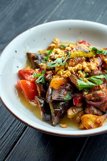 Appetizing, dietary salad of baked vegetables (tomatoes, peppers, eggplants) with sauce and nuts in a white plate on a dark wooden surface. Vegetarian food. Selective focus