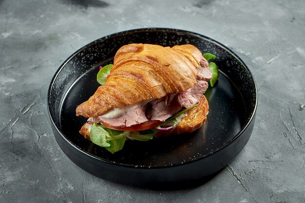 Appetizing French croissant sandwich with roast beef, tomatoes, spinach and white sauce, served in a black plate on a gray surface