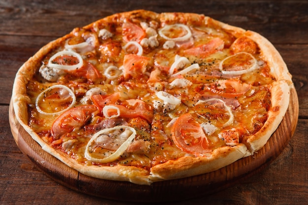 木製のテーブル、クローズアップで提供されるハム、タマネギ、トマト、チーズと食欲をそそる新鮮なおいしいピザ。全国のイタリア料理、レストランのメニュー写真。