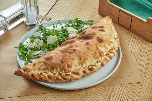 Appetizing freshly baked calzone pizza with crisp in a white plate on a wooden table. Restaurant serving.