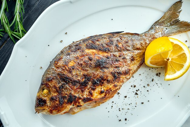 Appetizing grilled dorado on charcoal with lemon, served in a white plate on a dark wood surface. Seafood BBQ