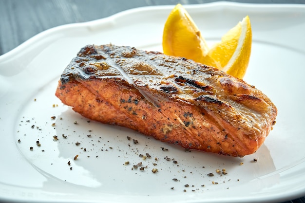 Appetizing grilled salmon steak on charcoal with lemon, served in a white plate on a dark wooden surface. BBQ seafood