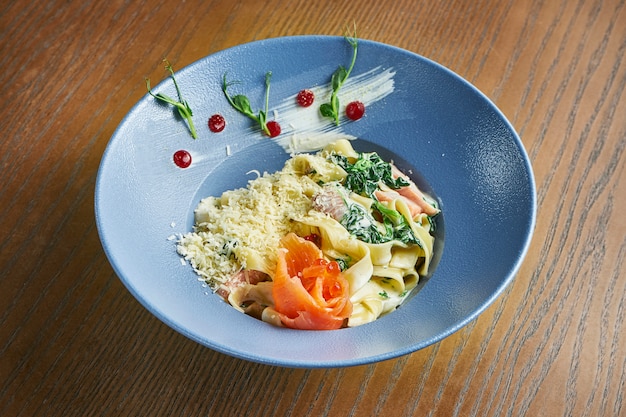 Appetizing, home-made tagliatelle pasta with spinach and salmon, parmesan in a blue bowl on a wooden surface. Italian cuisine. Add noise on post. Selective focus