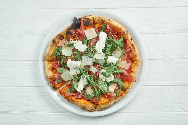 Appetizing homemade pizza with jamon, parmesan, arugula and straccella cheese on a white plate on a white